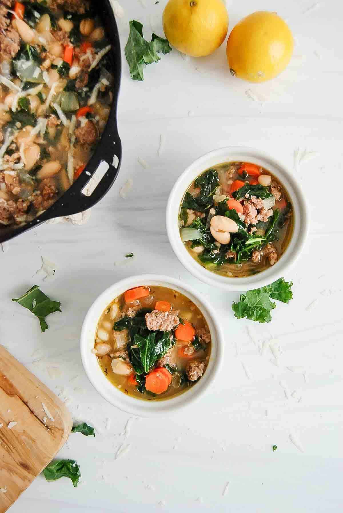 two bowls of sausage, kale and white bean soup with larger pot on the side.