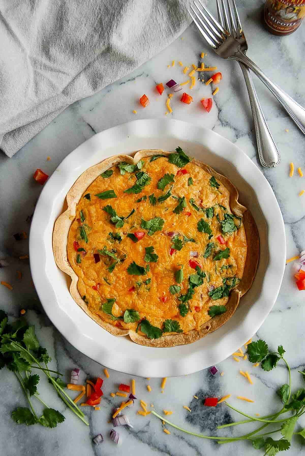pie plate with tortilla quiche bake on countertop with parsley on top and forks and hot sauce on the side.