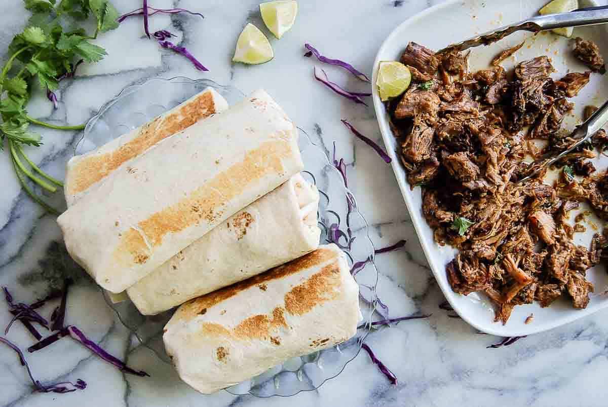 carnitas burritos on tray with plate of pork carnitas on the side.