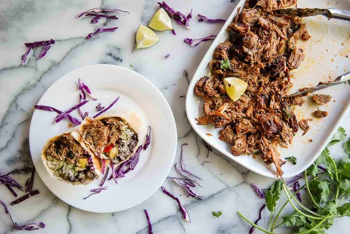 pork carnitas burrito cut in half on plate with full plate of carnitas on the side.