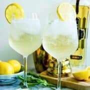 two wine glasses with hugo spritz, garnished with mint and lemon wheel with St-Germain bottle in the background.