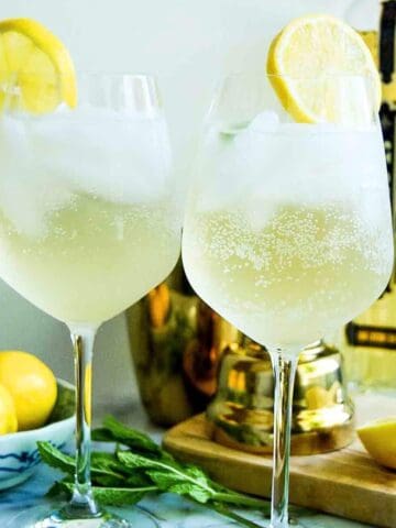 two wine glasses with hugo spritz, garnished with mint and lemon wheel with St-Germain bottle in the background.