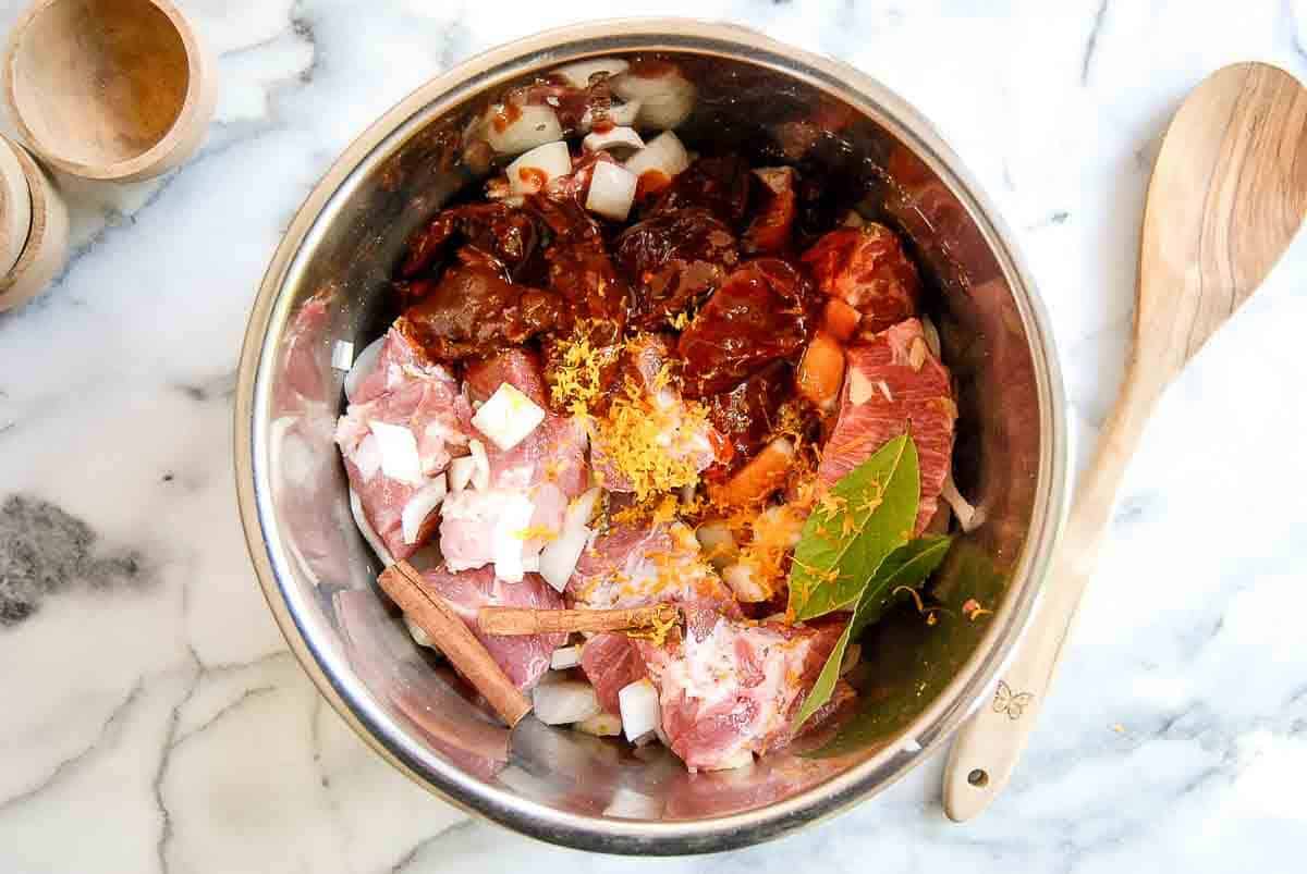 pork, onions, garlic, chipotle chiles, orange zest, bay leaves, cinnamon sticks orange juice, and spices in the instant pot with wooden spoon and bowls on the side.