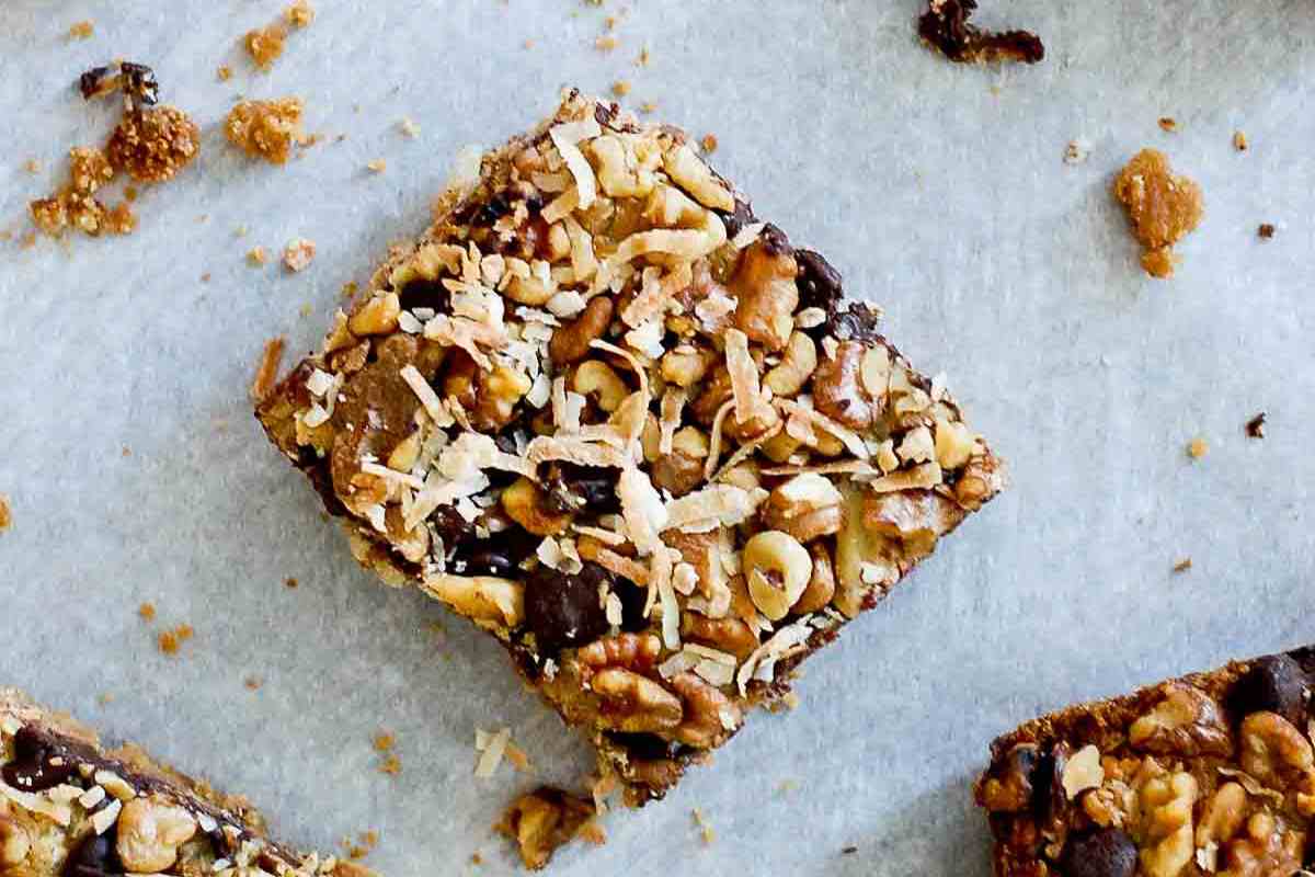 A close-up of a dessert bar topped with chopped nuts, chocolate chips, and shredded coconut, resting on parchment paper with a few crumbs scattered around.