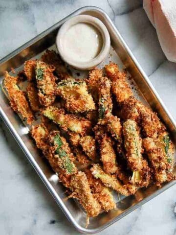 A metal tray filled with crispy breaded zucchini fries, served with a small cup of creamy dipping sauce, sits on a marble surface next to a folded pink towel.