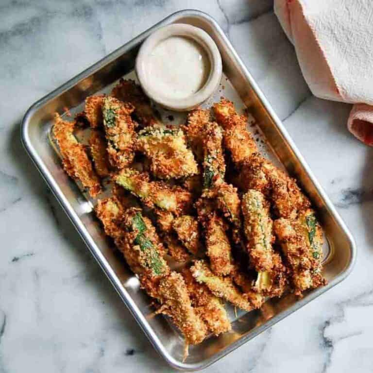 A metal tray filled with crispy breaded zucchini fries, served with a small cup of creamy dipping sauce, sits on a marble surface next to a folded pink towel.