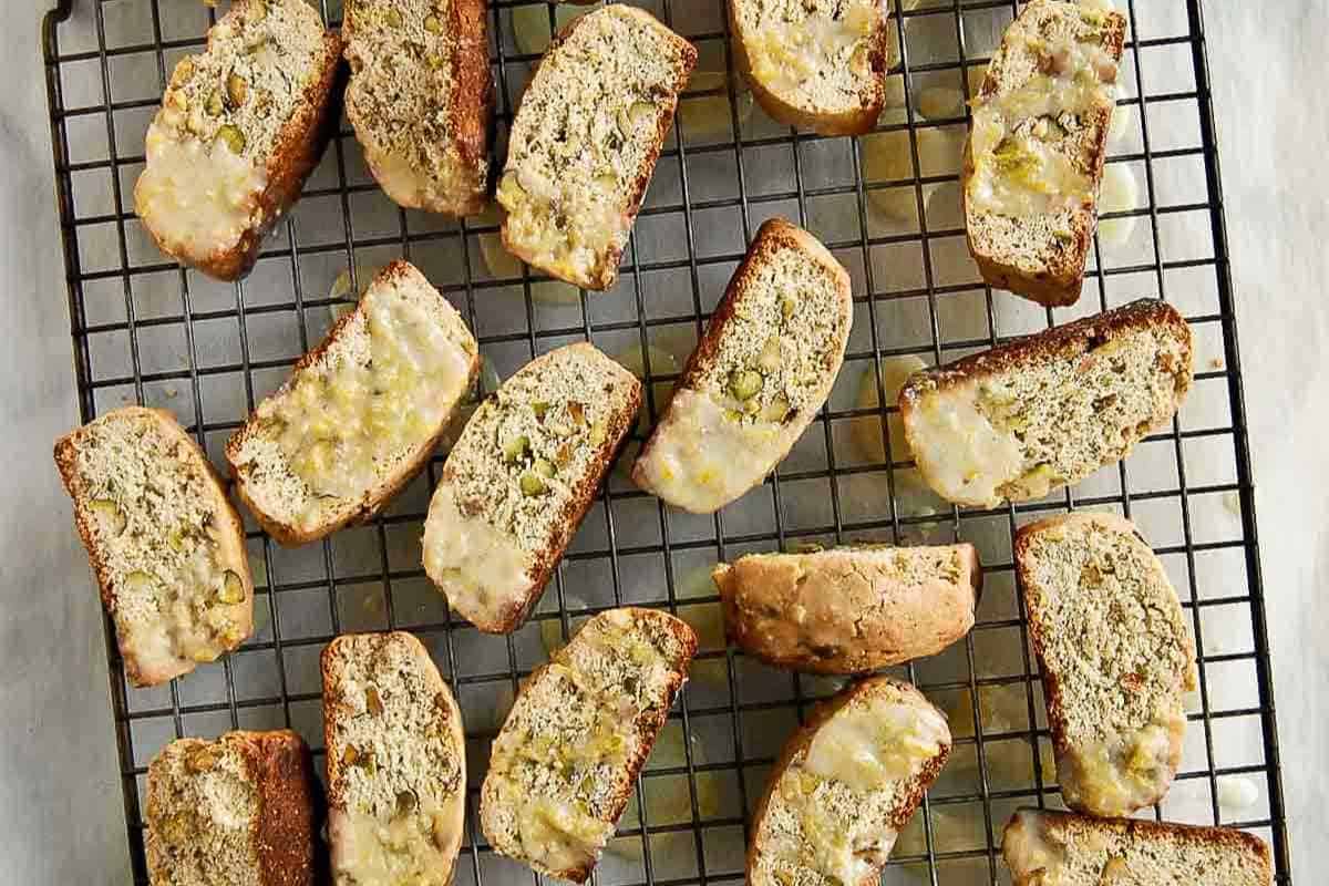 Rows of biscotti cookies with icing on top are arranged on a wire cooling rack. The cookies are golden brown and studded with nuts, and the icing appears to have dripped slightly over the edges.