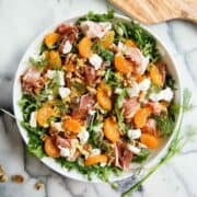 A salad in a white bowl with arugula, prosciutto, mandarin orange segments, walnuts, dollops of goat cheese, and fresh dill, on a marble surface with a wooden board partially visible.