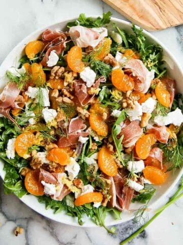 A salad in a white bowl with arugula, prosciutto, mandarin orange segments, walnuts, dollops of goat cheese, and fresh dill, on a marble surface with a wooden board partially visible.