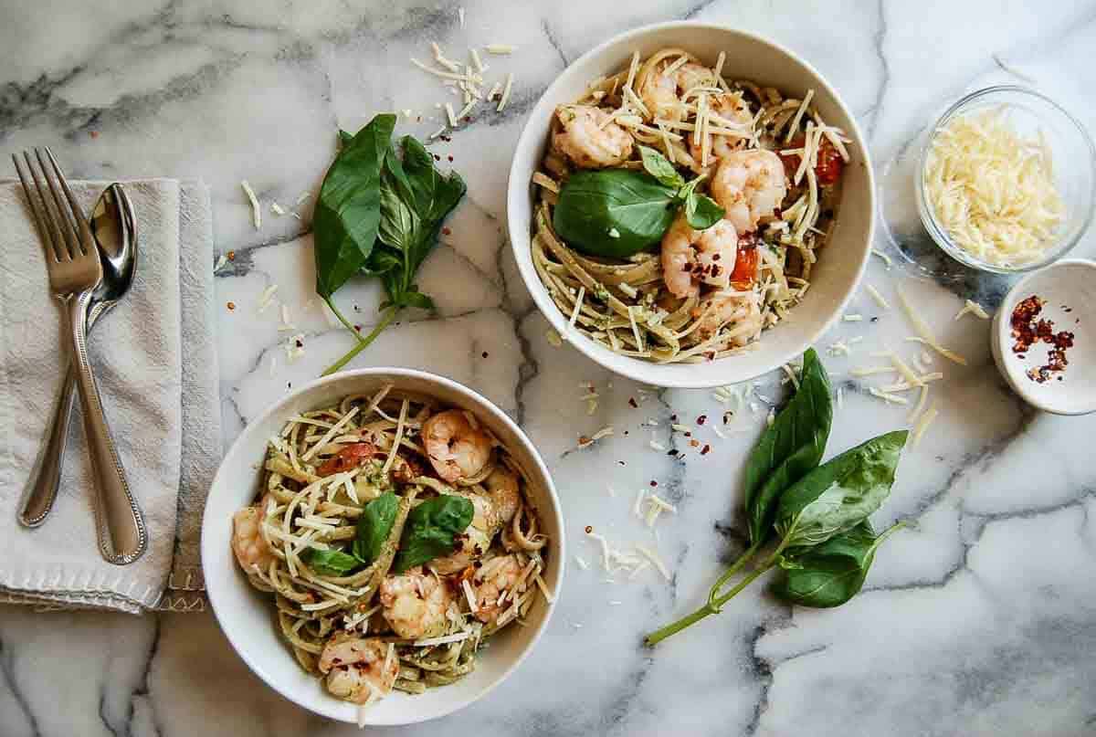 shrimp pesto pasta in two bowls on countertop with parmesan, basil and red pepper sprinkled on top.