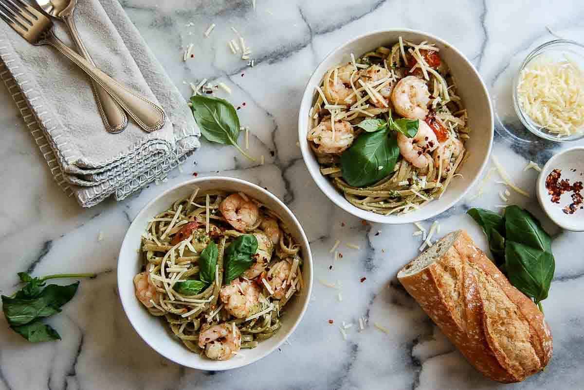 pesto shrimp pasta in two bowls on countertop with parmesan, basil and red pepper sprinkled on top.