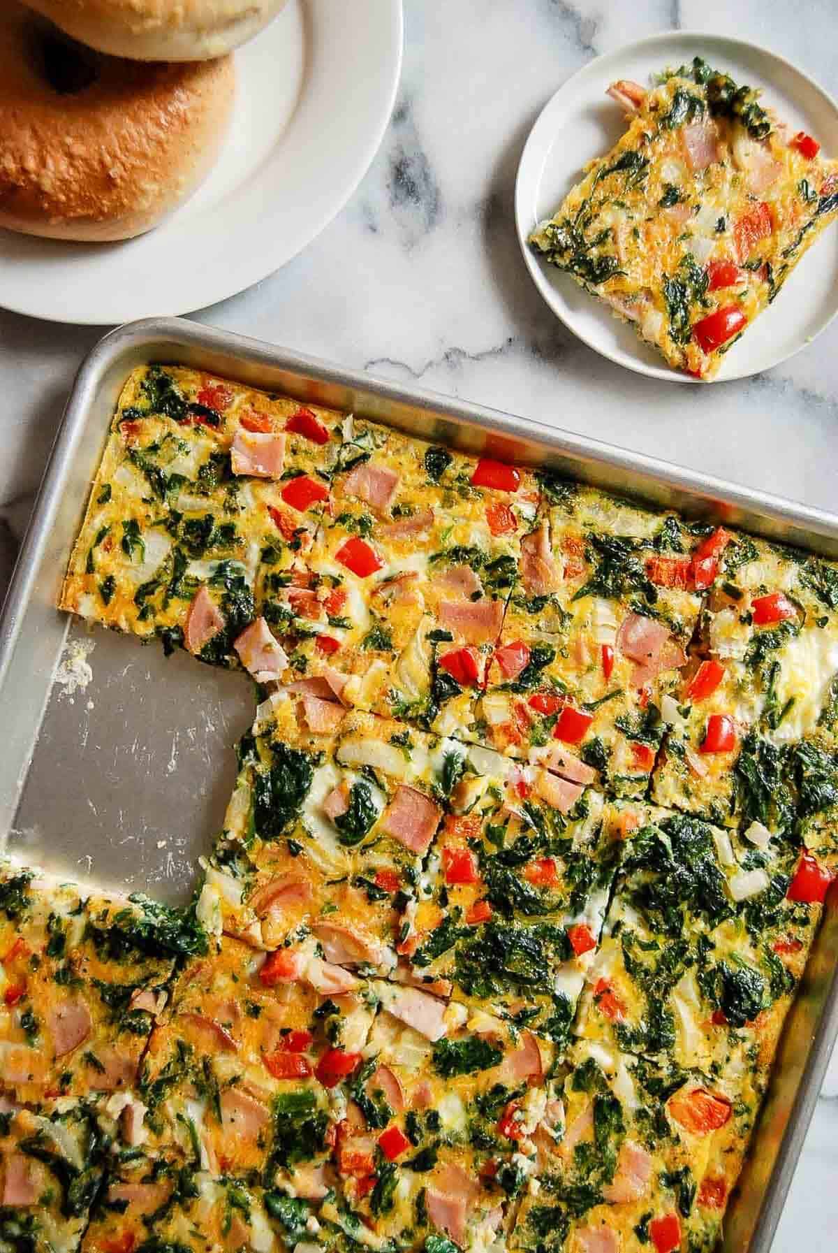 A sheet pan filled with baked egg squares containing spinach, red bell pepper, and ham sits on a marble surface. One square is served on a small plate, and a plate of bagels is nearby.