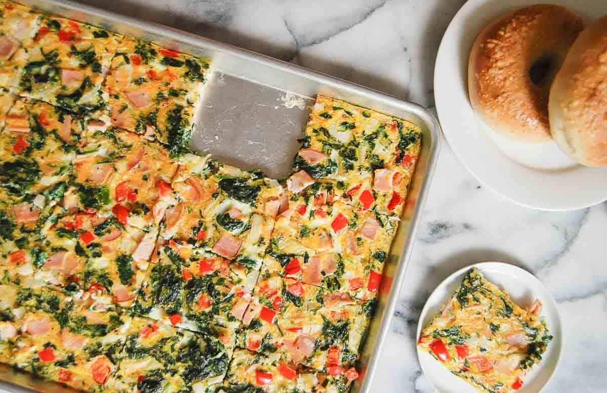 A sheet pan filled with baked egg squares featuring spinach, diced ham, and red bell pepper sits on a marble surface. Nearby are a plate of two bagels and a plate with a single egg square.