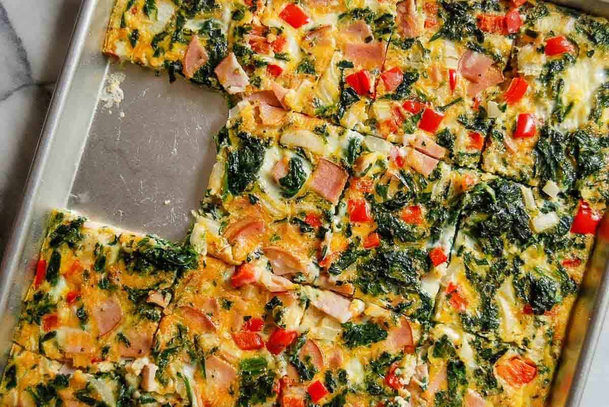 A sheet pan filled with a baked egg dish cut into squares. The dish contains visible pieces of ham, spinach, red bell pepper, and cheese. One square has been removed, revealing the baking pan underneath.