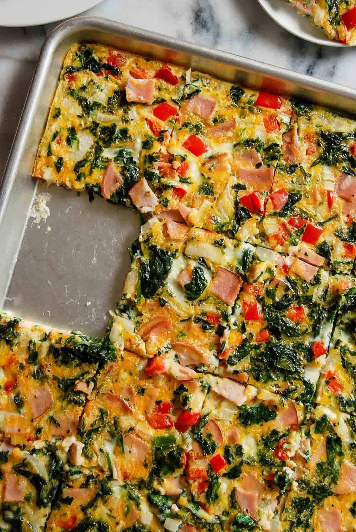 A sheet pan filled with baked egg squares topped with diced ham, spinach, and red bell peppers. Several squares have been cut out and one is missing from the tray.