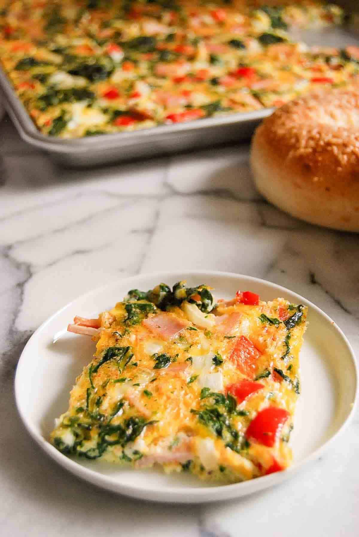 A slice of baked egg casserole with spinach, red peppers, and cheese on a white plate, with a whole casserole tray and a round bread roll in the background on a marble surface.