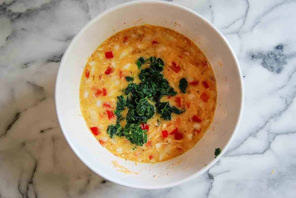 A white bowl on a marble surface filled with a mixture of eggs, diced red bell peppers, onions, and a garnish of chopped spinach on top.