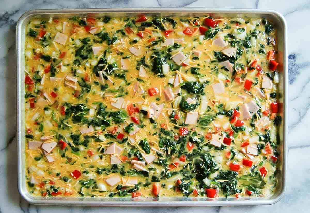 A sheet pan filled with a colorful egg mixture, including chopped spinach, red bell peppers, cheese, and small pieces of cooked meat, ready to be baked. The pan sits on a marble surface.