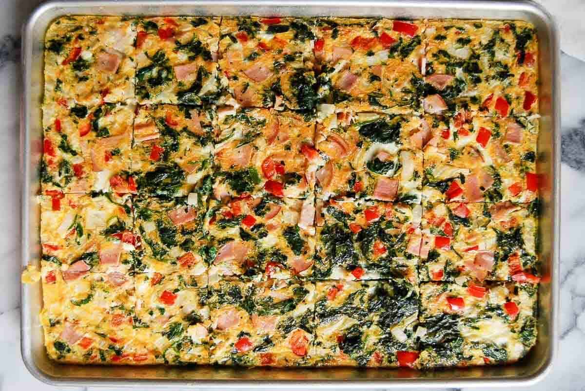 A large sheet pan filled with baked egg squares containing spinach, red bell peppers, and diced ham, cut evenly into portions on a marble surface.