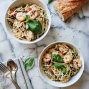 pesto shrimp pasta in two bowls on countertop with parmesan, basil and red pepper sprinkled on top.
