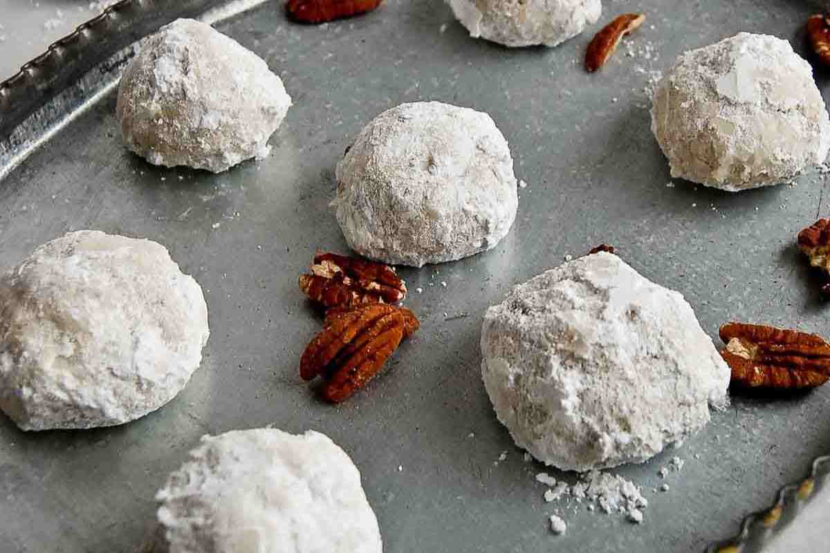 Powdered sugar-coated cookies and pecan halves are arranged on a metal baking tray.