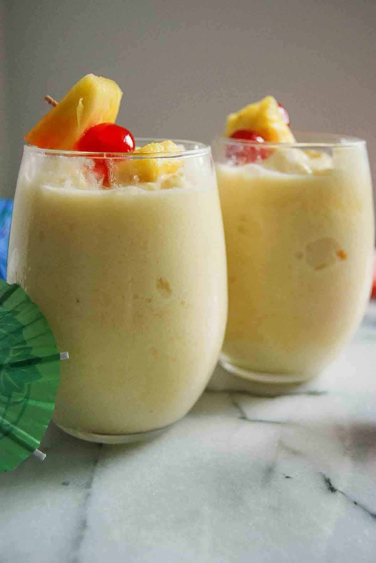 Two glasses of creamy yellow tropical drink garnished with pineapple chunks and red cherries, placed on a marble surface with a green paper umbrella nearby.