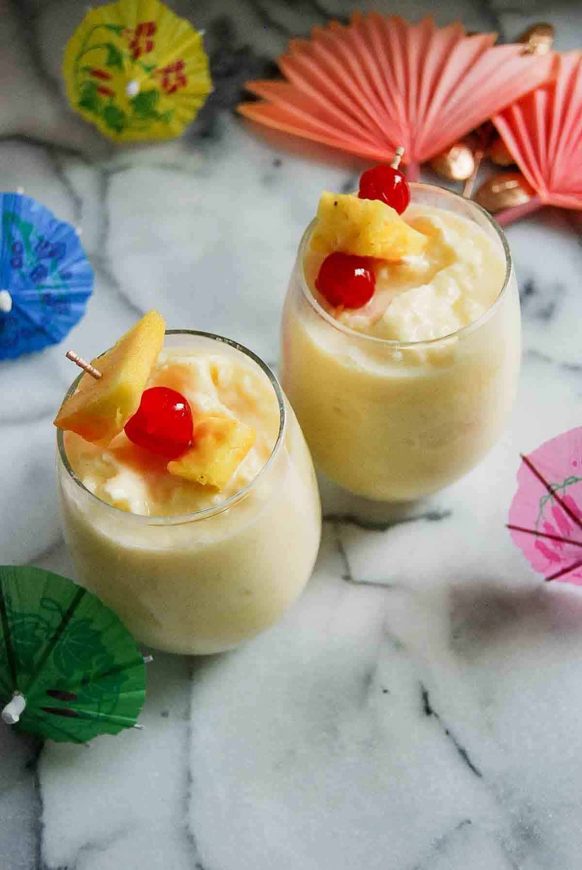 Two glasses of creamy yellow pina colada garnished with pineapple slices and maraschino cherries, surrounded by colorful paper cocktail umbrellas on a marble surface.