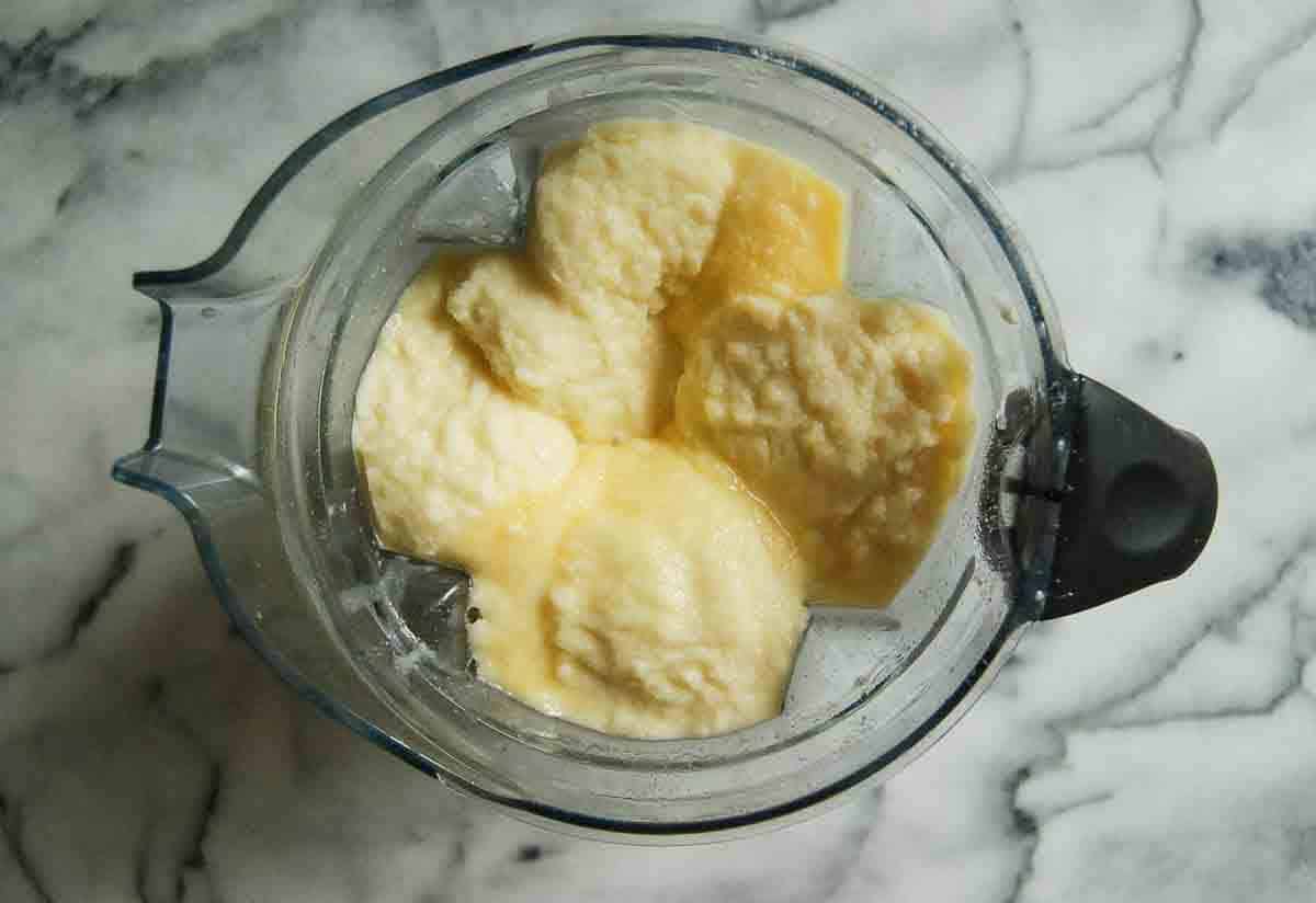 A blender filled with a thick, pale yellow mixture on a marble countertop, viewed from above.