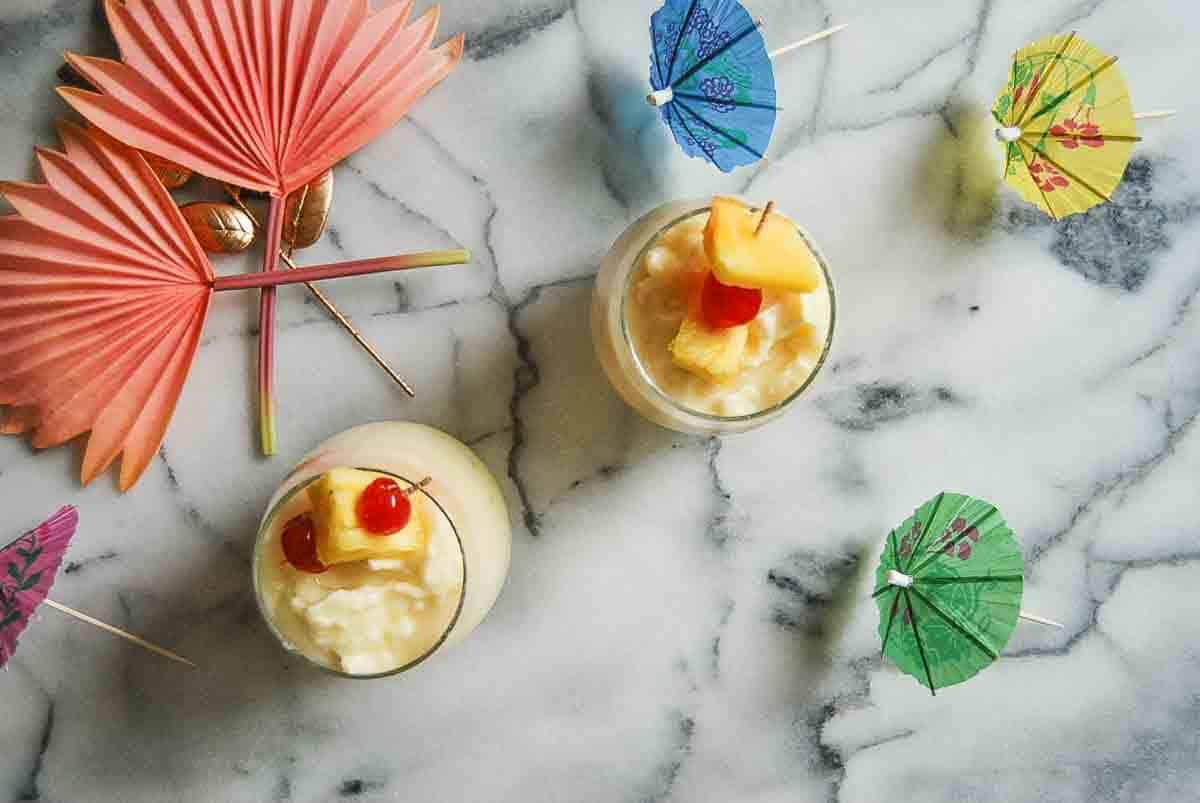 Two glasses of creamy tropical drinks topped with fruit and cherries sit on a marble surface, surrounded by colorful paper umbrellas and fans, creating a summery, festive vibe.