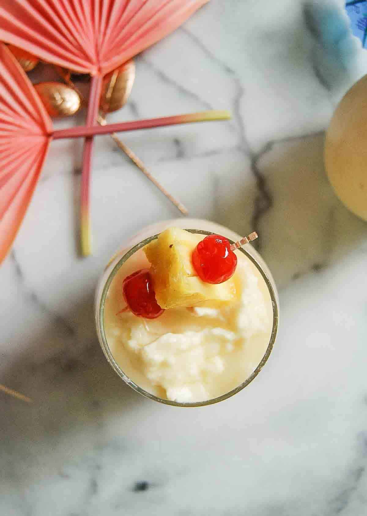 A creamy tropical drink garnished with a pineapple chunk and two maraschino cherries, served in a glass on a marble surface with pink cocktail umbrellas nearby.