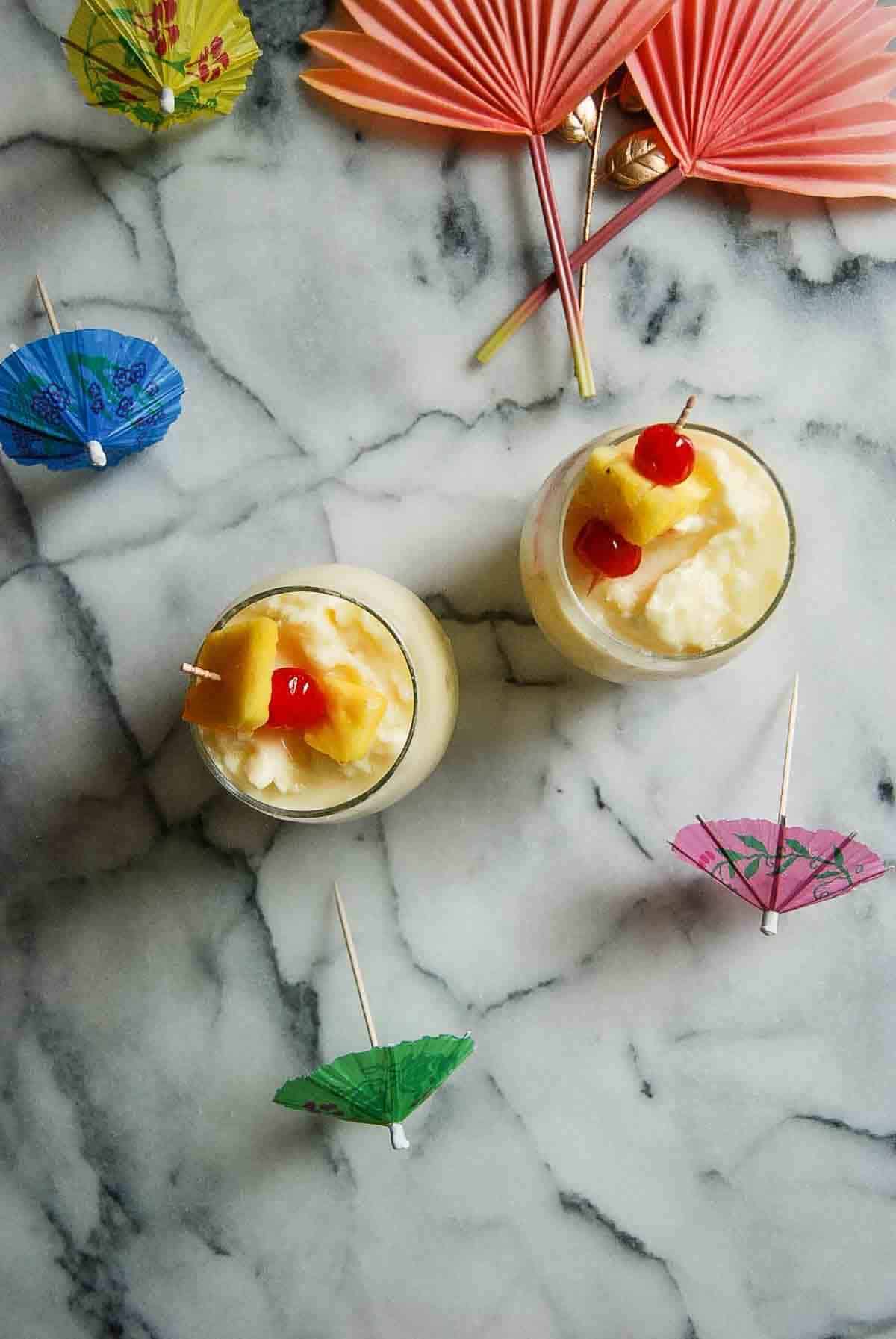 Two tropical cocktails with pineapple and cherry garnishes are on a marble surface, surrounded by colorful paper umbrellas and two pink folding fans.