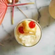 A tropical drink topped with a pineapple chunk and two maraschino cherries, garnished with a cocktail umbrella, viewed from above on a marble surface.