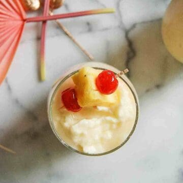 A tropical drink topped with a pineapple chunk and two maraschino cherries, garnished with a cocktail umbrella, viewed from above on a marble surface.