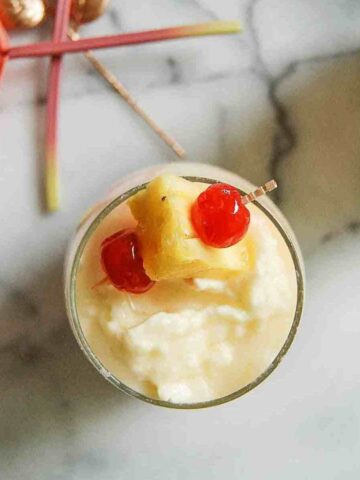 A tropical drink topped with a pineapple chunk and two maraschino cherries, garnished with a cocktail umbrella, viewed from above on a marble surface.