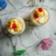 Two glasses of creamy tropical drinks topped with pineapple chunks and cherries, viewed from above on a marble surface, surrounded by colorful paper drink umbrellas.
