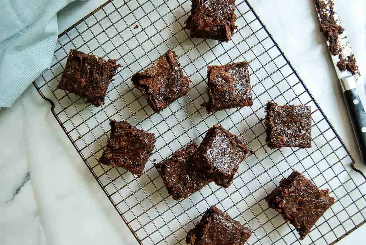 Nine chocolate brownies are arranged on a cooling rack over a white marble surface, with a knife and a folded light blue cloth in the corners of the image.