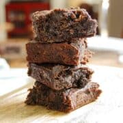 A stack of four thick, fudgy brownies is piled on a wooden surface, with a blurred background of a home interior. The brownies appear rich, moist, and chocolatey with a slightly crumbly texture.