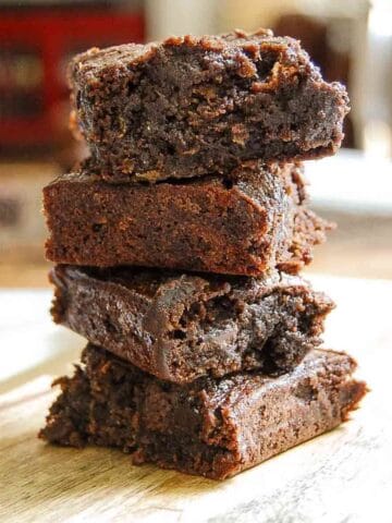 A stack of four thick, fudgy brownies is piled on a wooden surface, with a blurred background of a home interior. The brownies appear rich, moist, and chocolatey with a slightly crumbly texture.