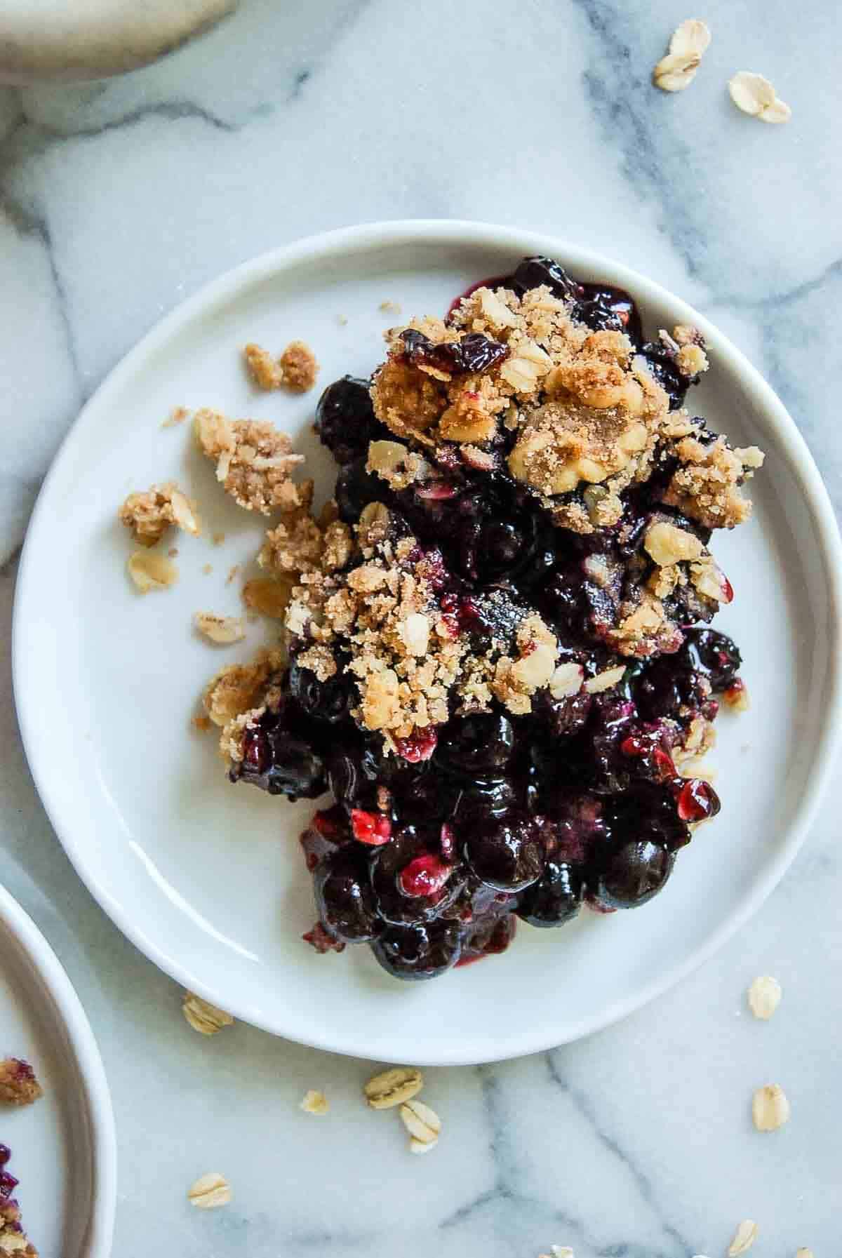 A serving of blueberry crisp with oat topping on a white plate, set on a marble surface. Some crumbs and oats are scattered around the plate.