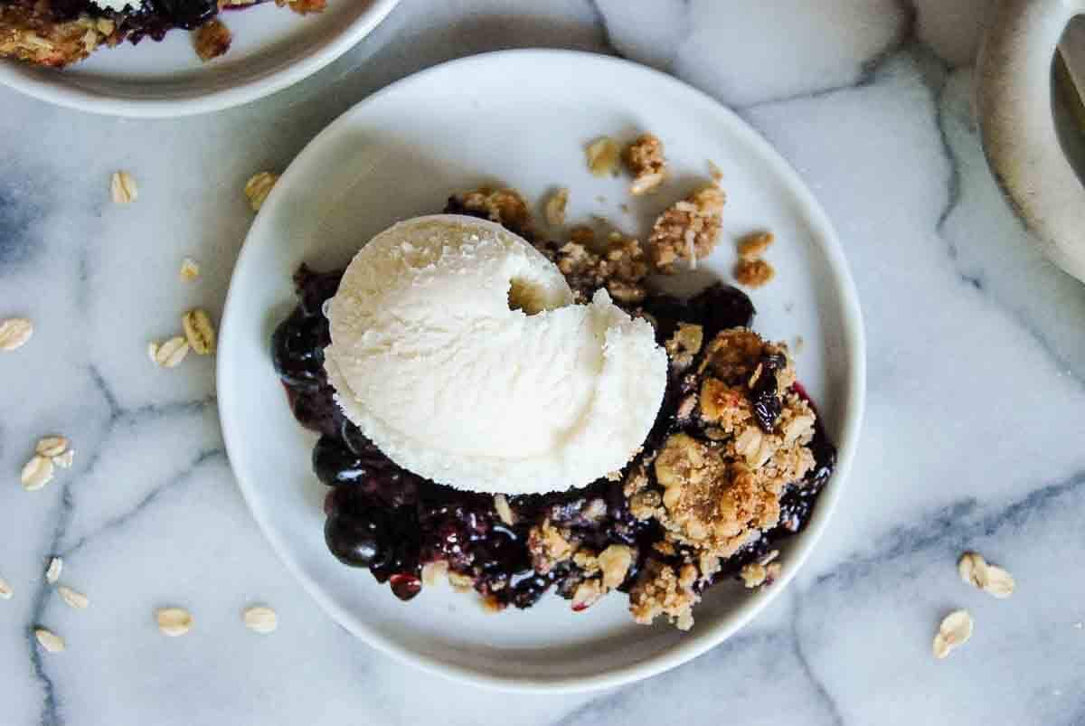 A white plate with a serving of berry crisp topped with a scoop of vanilla ice cream, placed on a marble surface with a few scattered oats around.