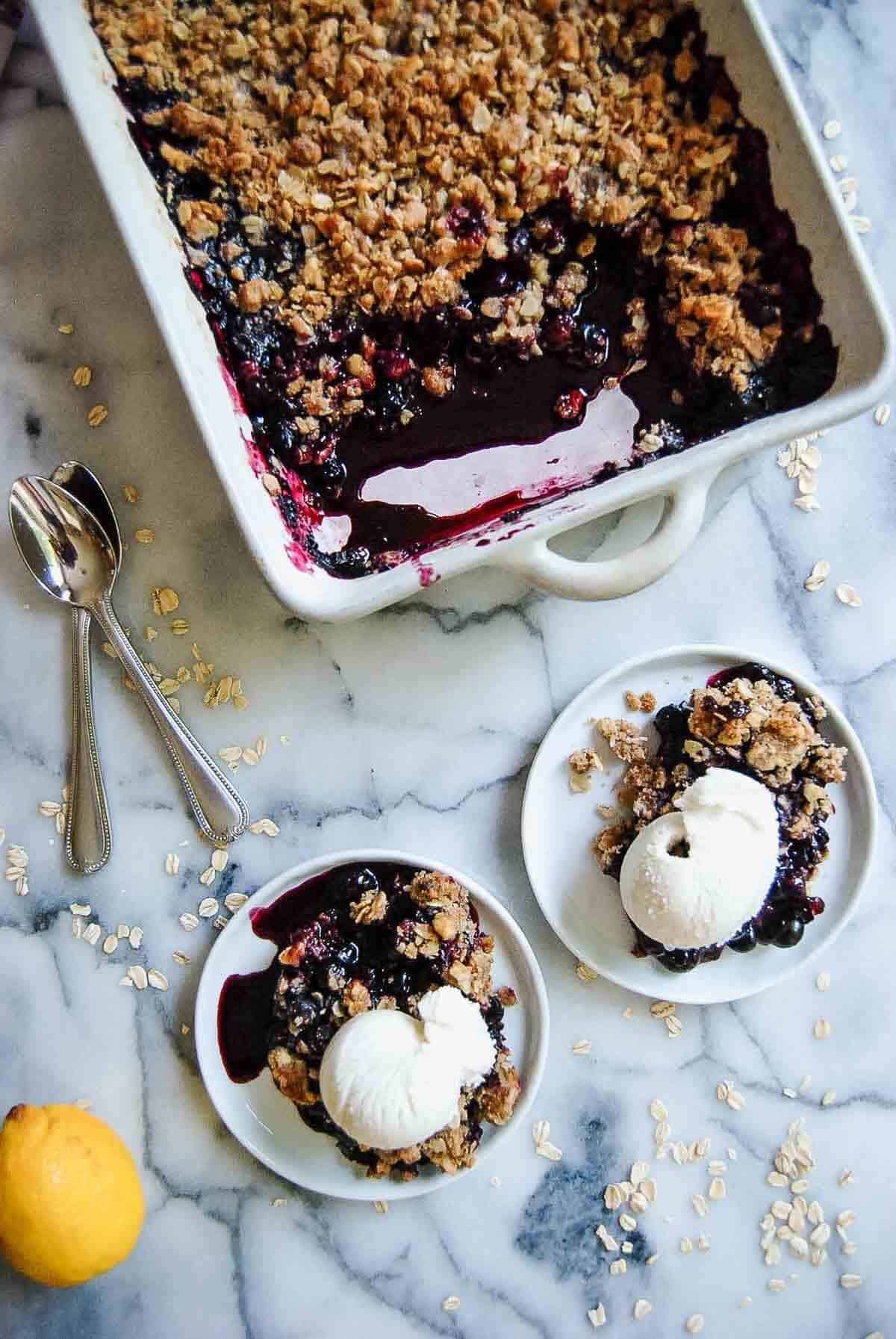 A baking dish of berry crisp sits on a marble counter with two plates of crumble topped with vanilla ice cream. Spoons, rolled oats, and a lemon are nearby, adding to the dessert scene.
