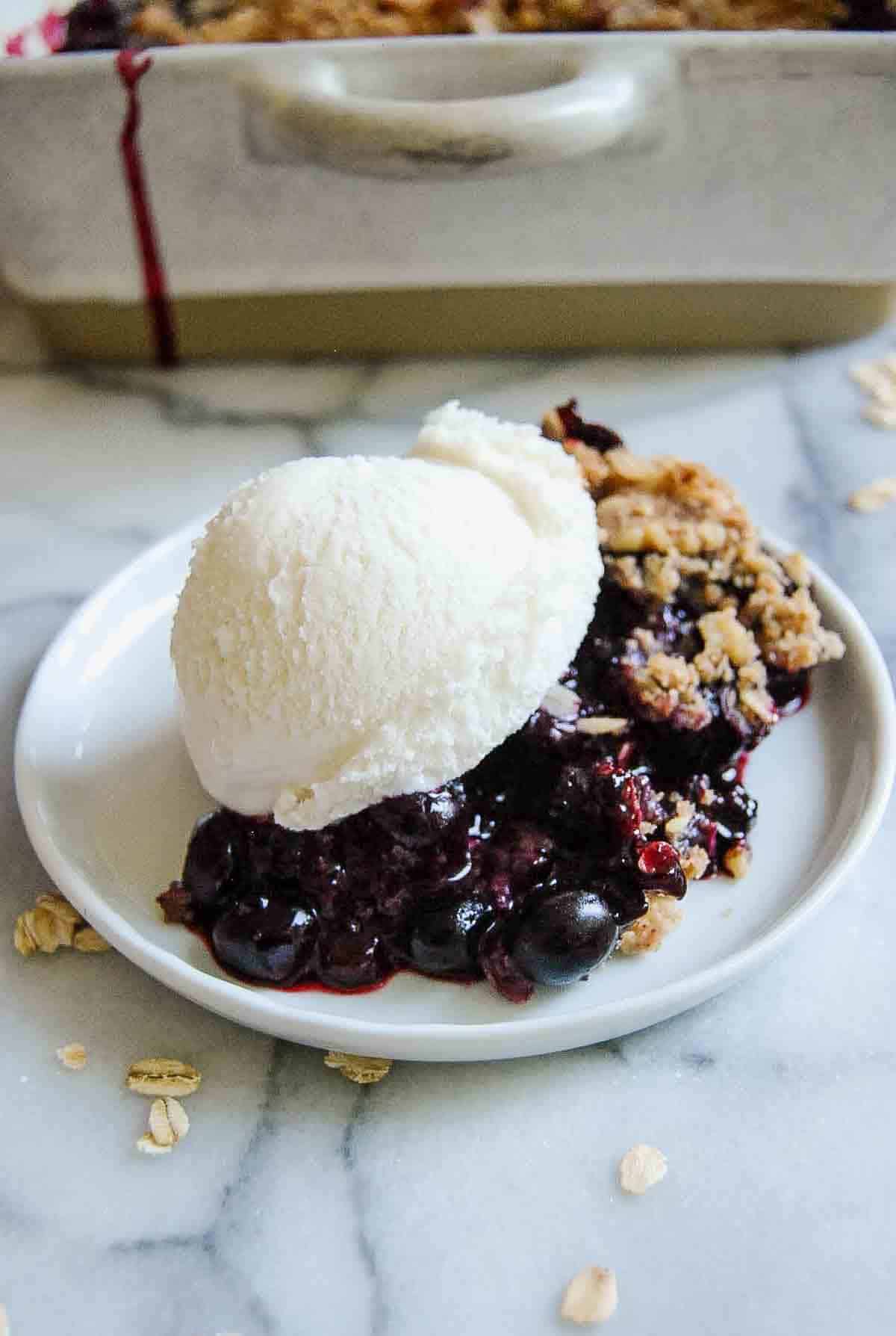 A serving of blueberry crisp with a scoop of vanilla ice cream on top, displayed on a white plate. The dessert features juicy blueberries and a crumbly oat topping.