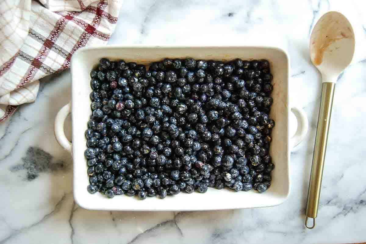 A white baking dish filled with fresh blueberries sits on a marble countertop next to a wooden spoon with batter and a checkered kitchen towel.