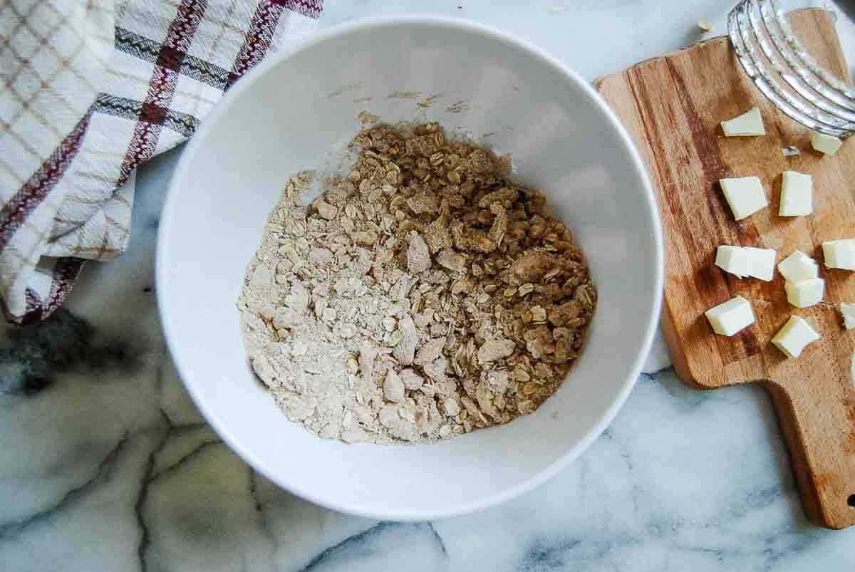 A white bowl with a mixture of flour and oats sits on a marble surface beside a wooden cutting board with small cubes of butter and a pastry cutter, with a plaid kitchen towel nearby.