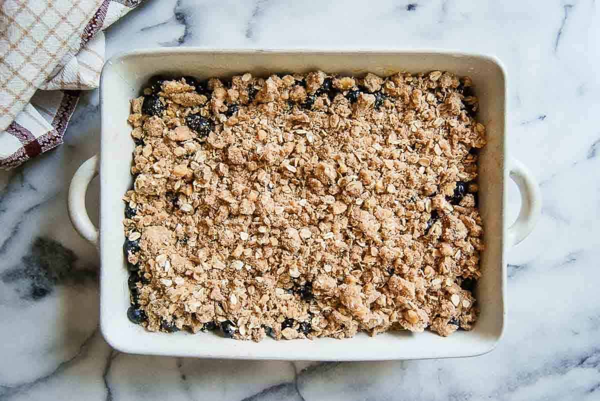 A rectangular white baking dish filled with blueberry crisp, topped with fresh oat topping, unbaked, sits on a marble countertop next to a folded dish towel.