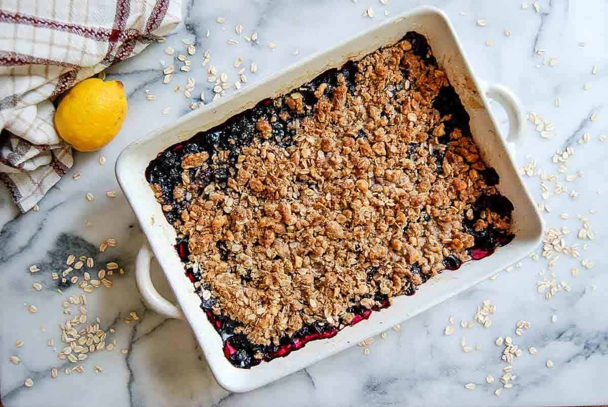 A baked blueberry crisp in a rectangular white dish sits on a marble surface, topped with a golden oat crumble. Scattered oats, a lemon, and a plaid towel are nearby.