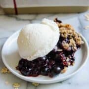 A white plate holding a serving of blueberry crisp topped with a scoop of vanilla ice cream, resting on a marble surface. Some oats are scattered nearby.