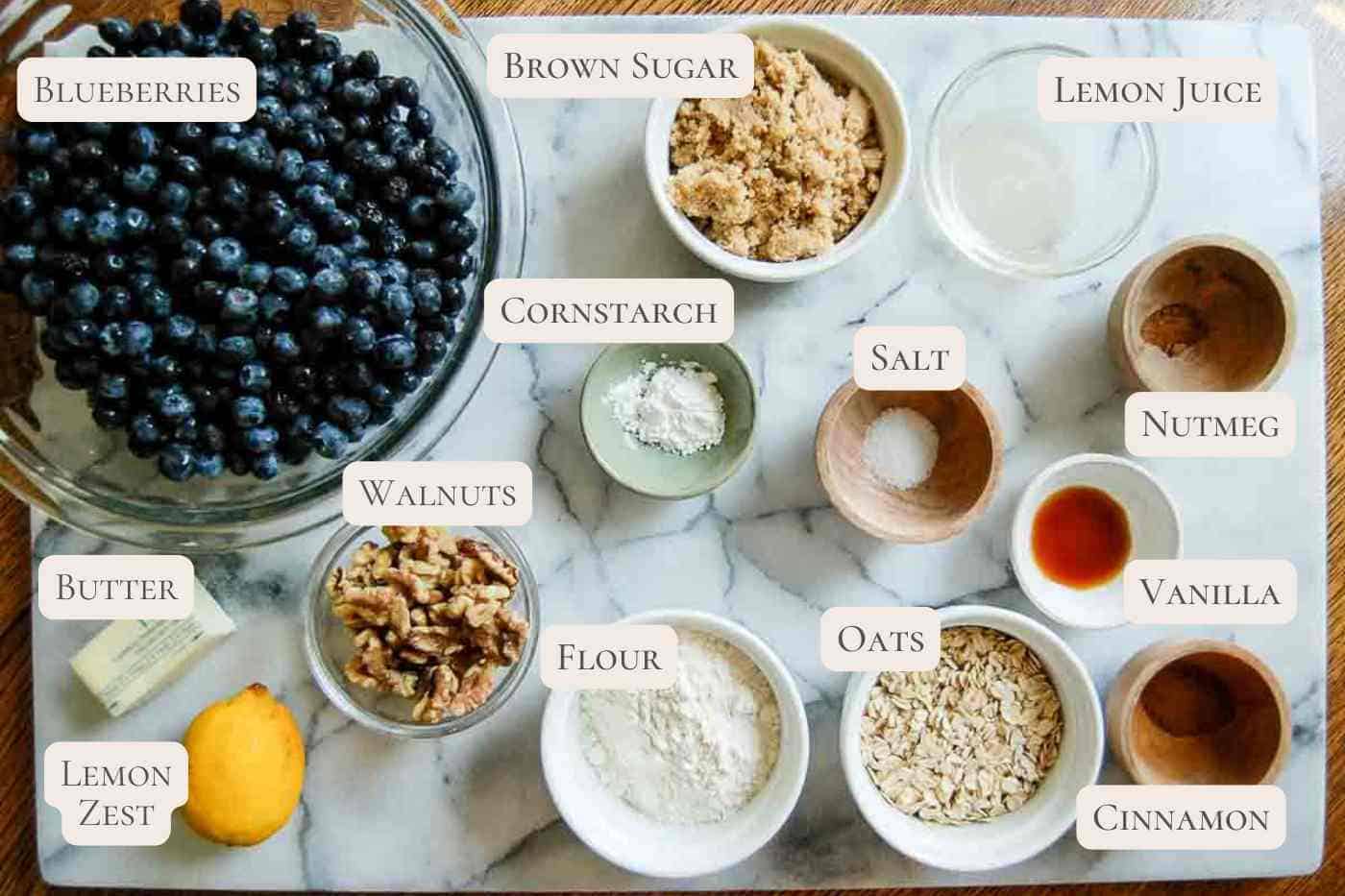 A marble surface displays labeled bowls of blueberries, brown sugar, lemon juice, cornstarch, salt, nutmeg, vanilla, cinnamon, oats, flour, walnuts, butter, and a lemon with lemon zest.