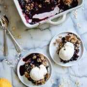 A baking dish of blueberry crisp sits on a marble countertop next to two small plates, each with a serving of crisp topped with vanilla ice cream. Nearby are two spoons, scattered oats, and a whole lemon.
