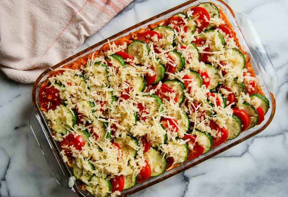 A glass baking dish filled with neatly arranged slices of zucchini and tomato, topped with shredded cheese and breadcrumbs, sits on a marble surface next to a pink kitchen towel.